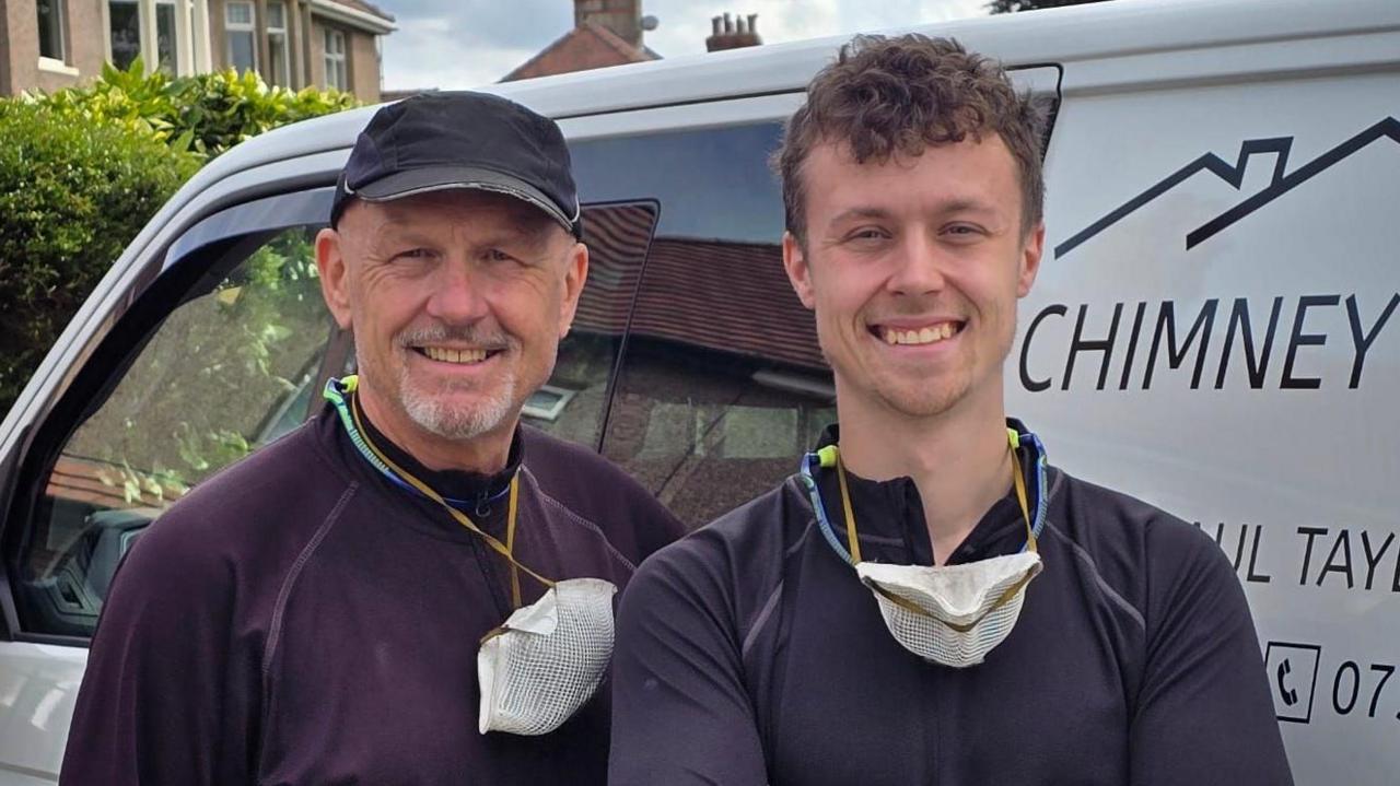 Father and son chimney sweeps Paul (left) and Matthew Taylor (right) standing in front of a white van wearing navy tops and with white masks around there necks. They are both smiling. 