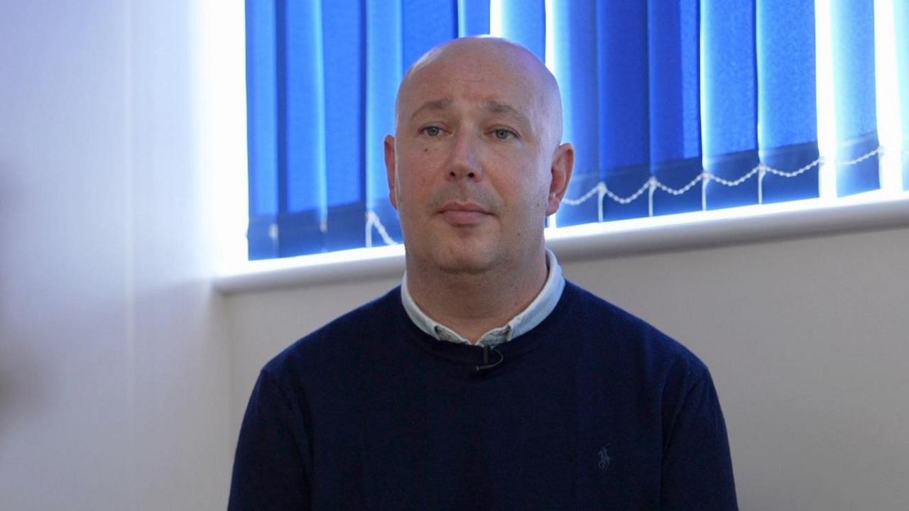 A man in a blue jumper sat next to a white wall and a window with a blue blind