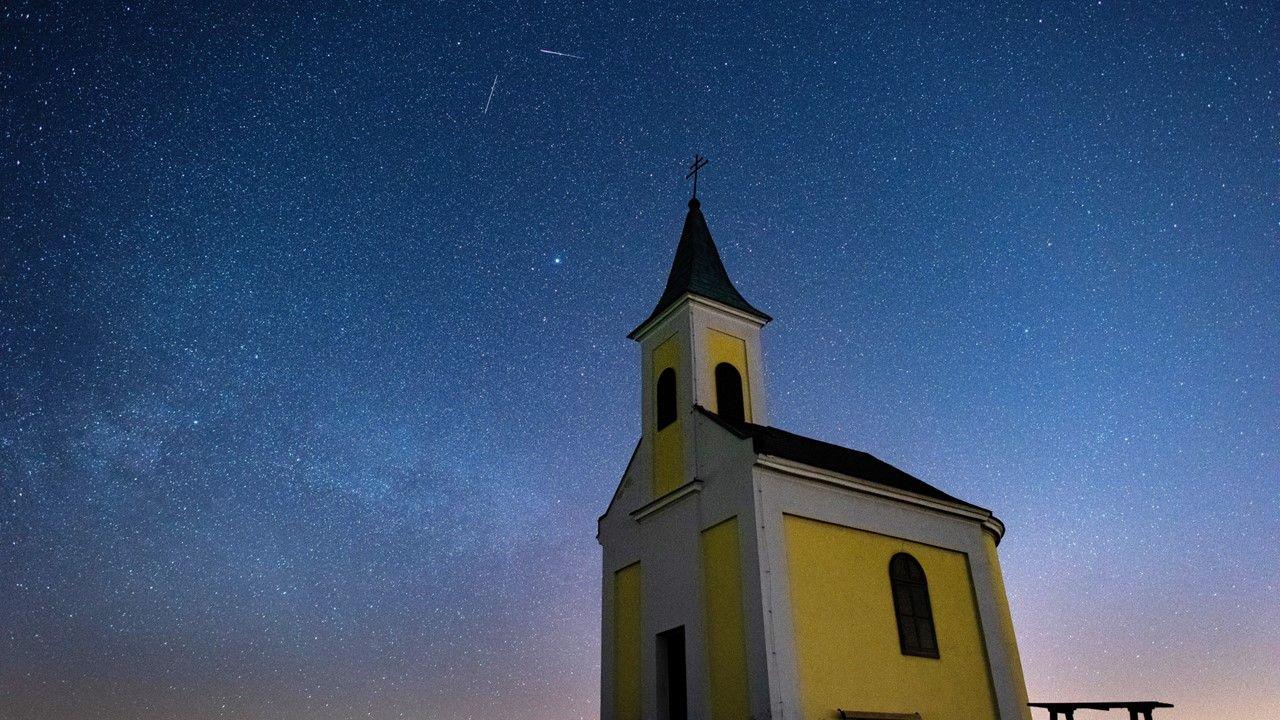 Clear lilac and dark blue night skies showing the Lyrids meteor shower streaks with a bench and yellow, grey and black church in foreground