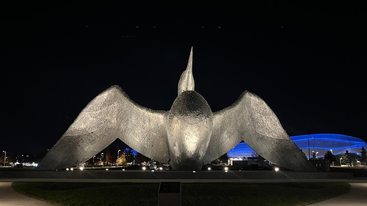 The Calling steel sculpture of a loon bird with its head thrown back and wings fully outstretched, installed on a paved/grass area with lights illuminating it from below