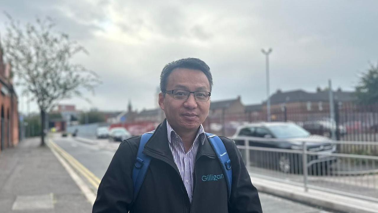 Colin Tang wearing a black coat with a purple and white stripped shirt beneath. He is wearing black glasses and has a blue backpack on his shoulders.