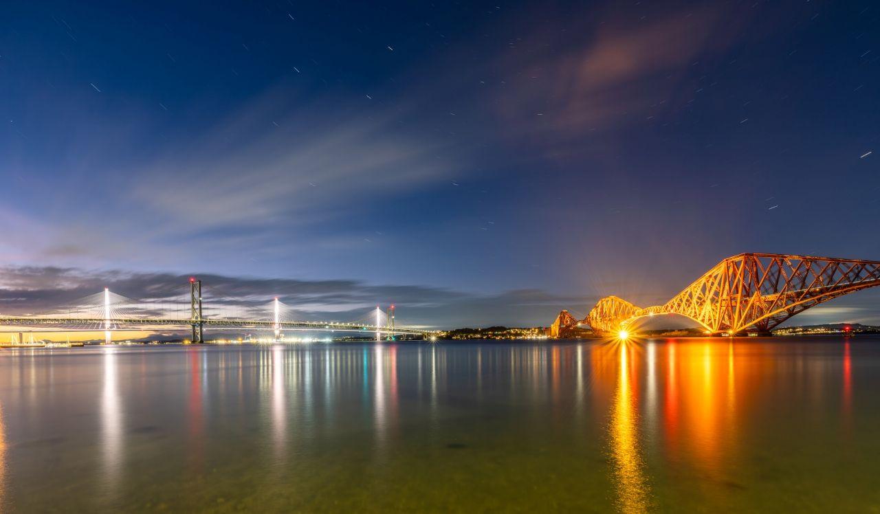The Forth Bridge and Queensferry Crossing taken with a long exposure. The bridges are lit up and the lights are reflected in the river