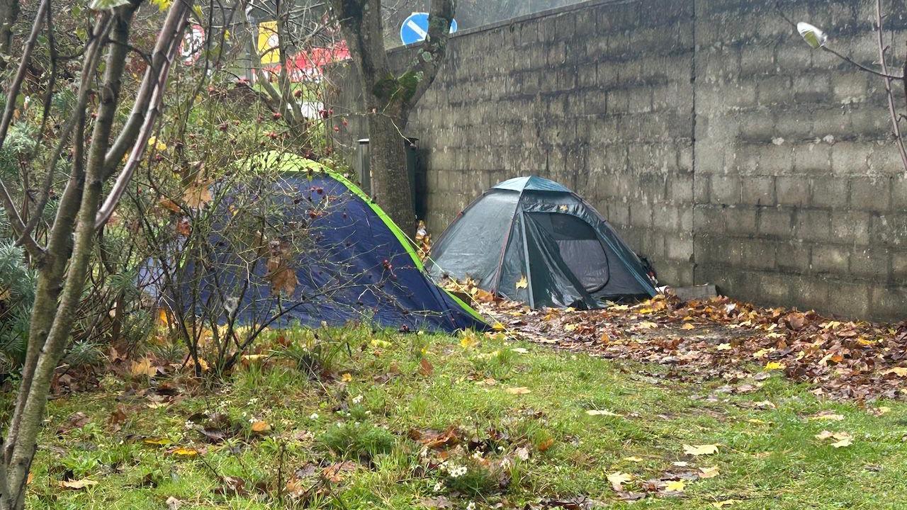 Two blue tents are seen on grass near a concrete retaining wall. The weather is drizzly. 