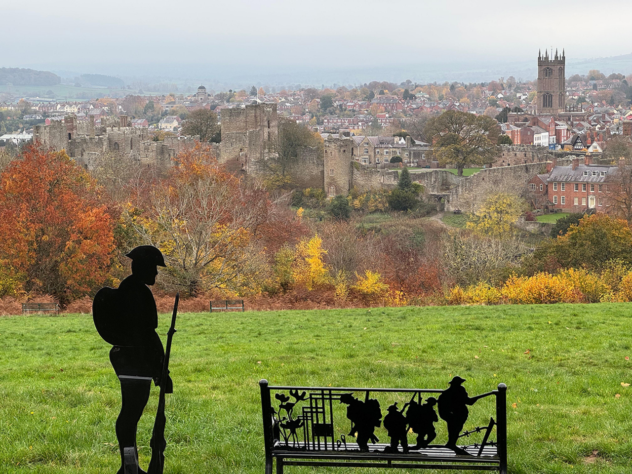 In the foreground is a metal sculpture of a soldier in a helmet holding a rifle on the ground. Next to him is a bench with its back carved into the shapes of four soldiers walking past barbed wire and with poppies to the left. The town of Ludlow is in the background, with the castle and St Laurence's Church particularly prominent