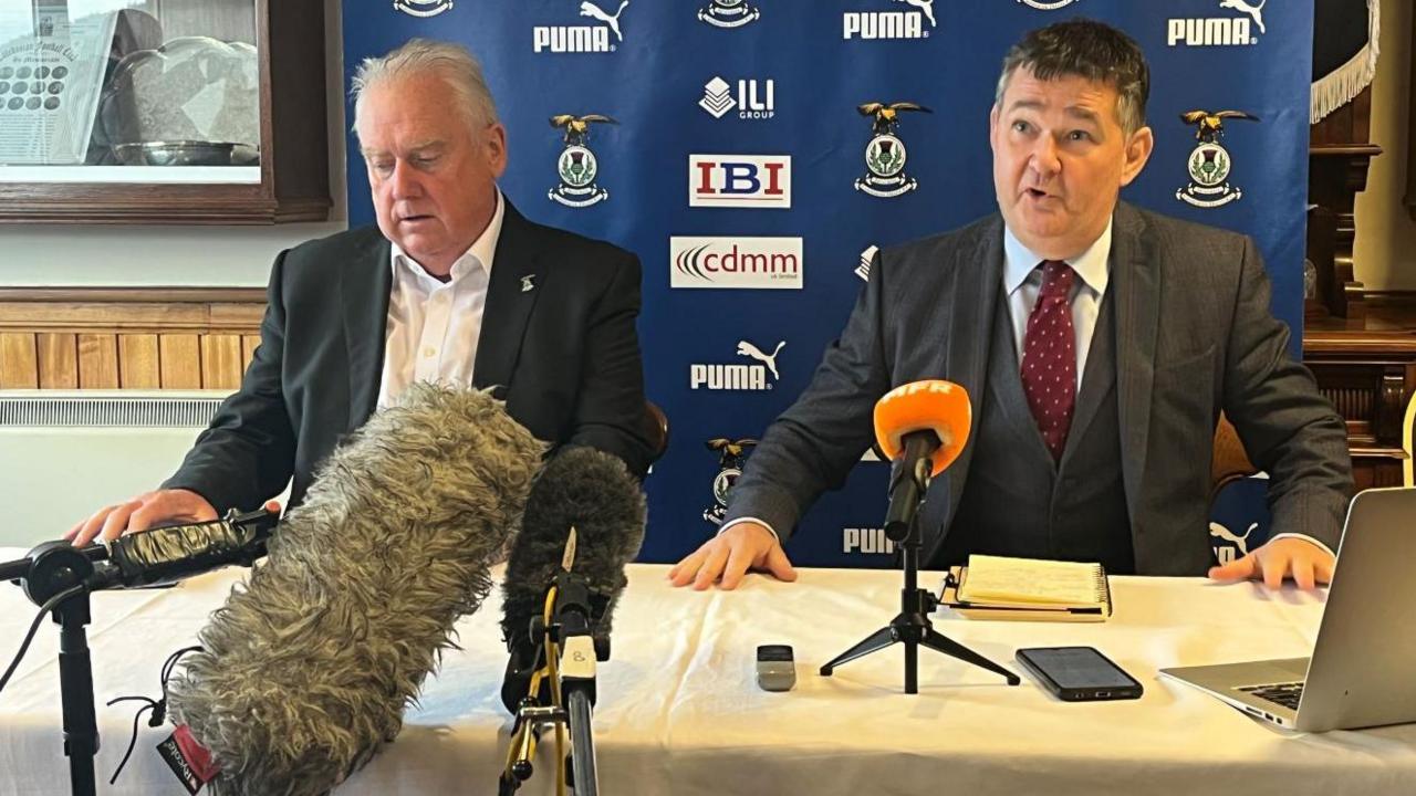 Alan Savage, left, and administrator James Stephen sit at a table. There are microphones and a laptop in front of them.