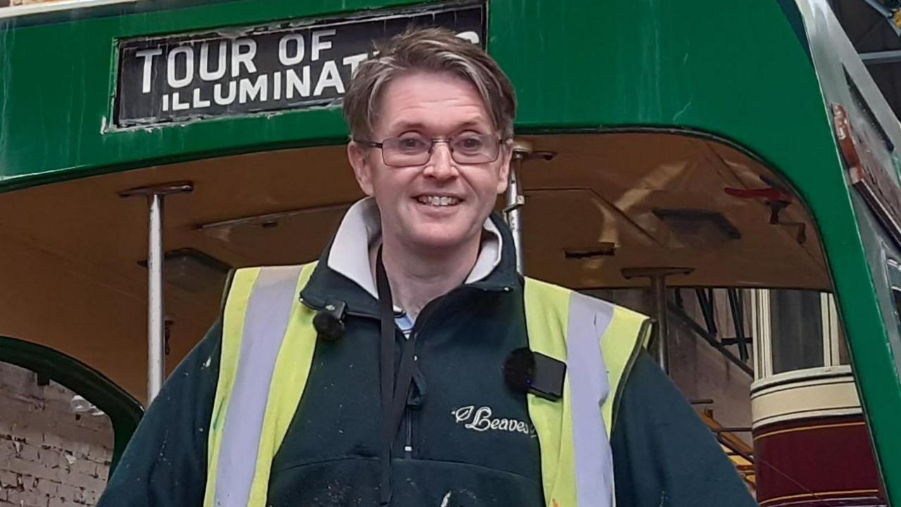 Councillor Paul Galley stands on a green-liveried tram in a workshop wearing a yellow high-vis vest over a black fleece. With dark, centre-parted hair and wearing glasses, he smiles for the camera.
