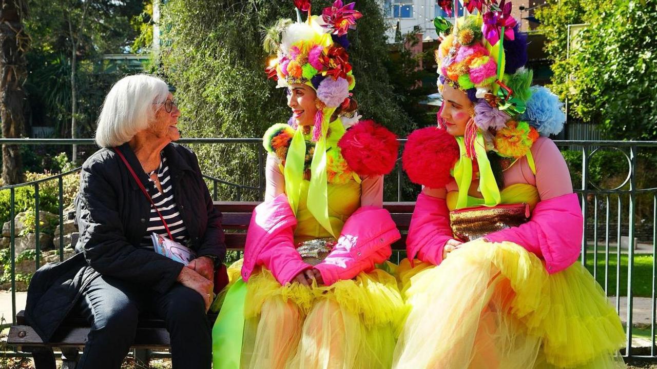 Two women in brightly coloured dresses sat on a bench with another woman who is not dressed in performative clothing. The group is chatting.