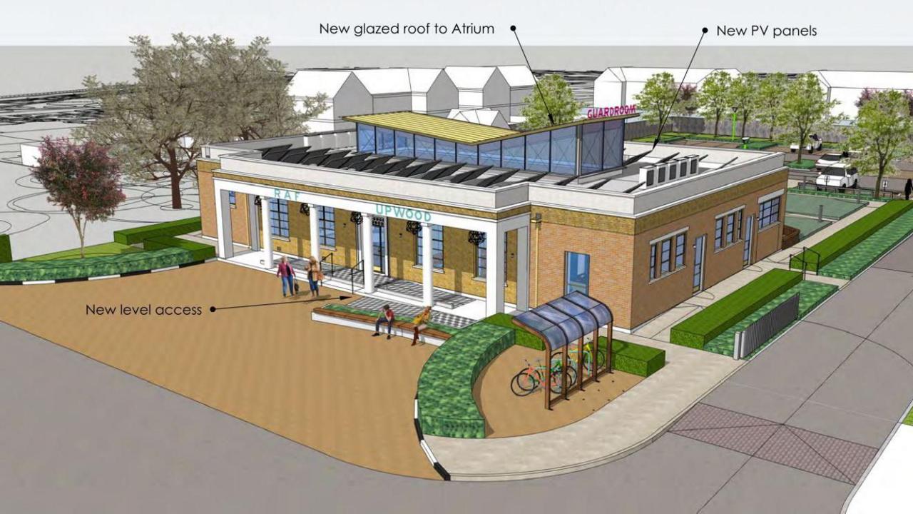 Single-storey building with white facade with six pillars equally spaced. There is a brick wall with windows behind the facade. The RAF Upwood legend above the entrance remains. There are windows to the side and a raised glass roof in the middle of the main roof. There is a covered cycle rack to the left of the front elevation. Trees and shrubs are visible around the building.