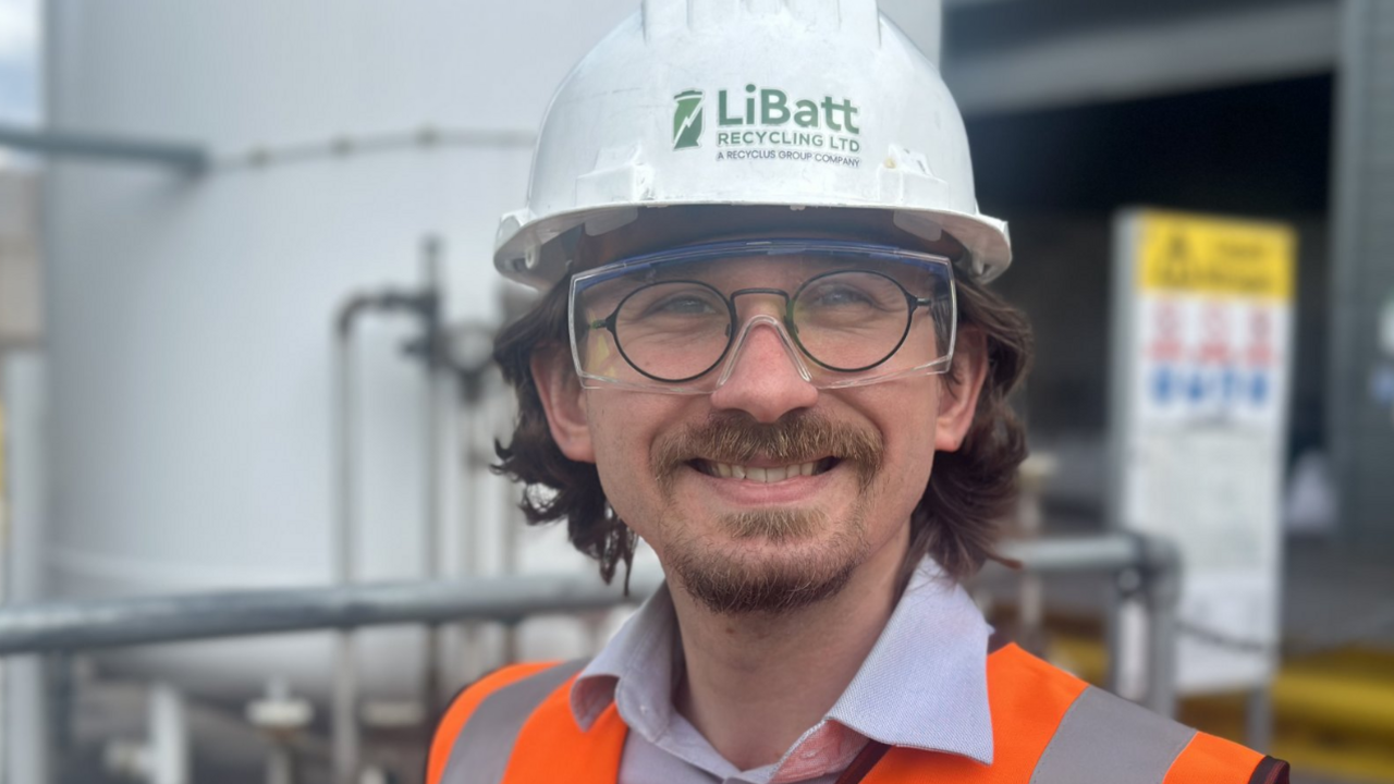 Dr Zorin is smiling at the camera and has glasses with safety goggles on over the top. He is wearing a safety helmet with a hi-vis bib on over a blue shirt. He has a beard and dark brown curly hair.