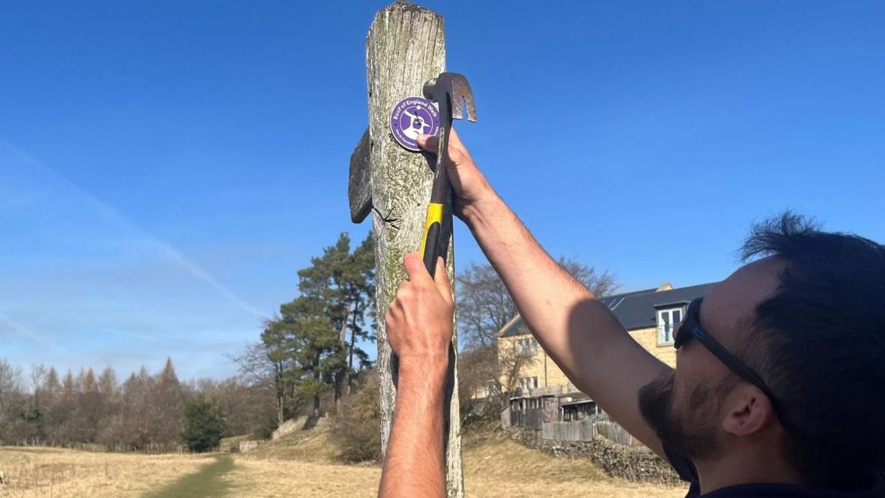 Side view of a man hammering a round purple plaque onto a post. He has dark hair and a beard and is wearing sunglasses. In the background is a rural view of a field and trees and to the right a partial view of a large house, all under blue skies.