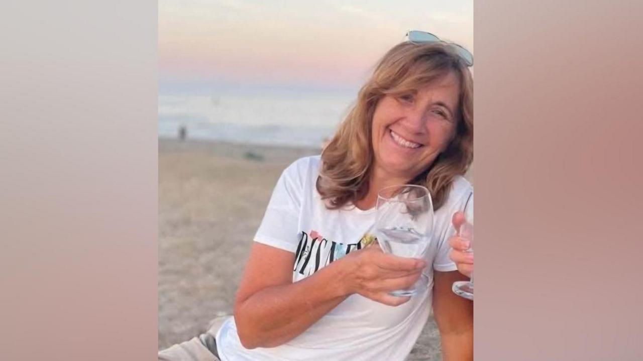 A woman wearing a white T-shirt smiling while holding a glass and sitting on a beach. 