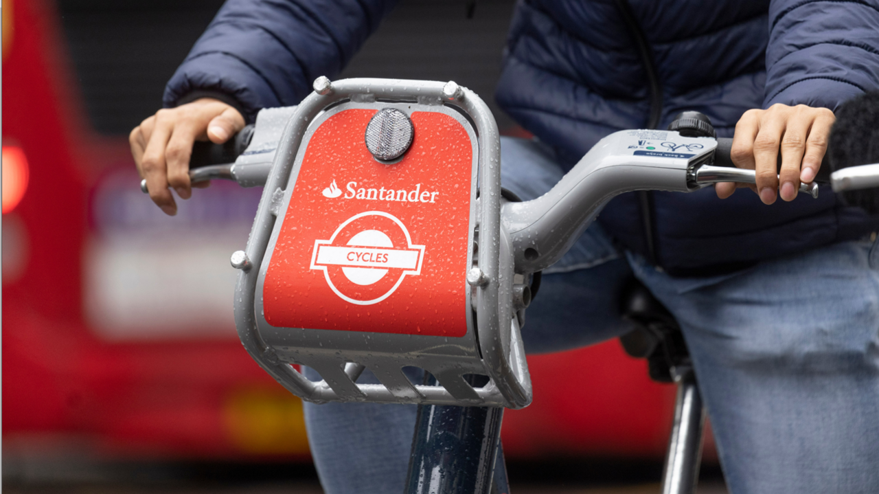 Red Santander branded e-bikes ridden by someone wearing jeans and a blue puffer jacket