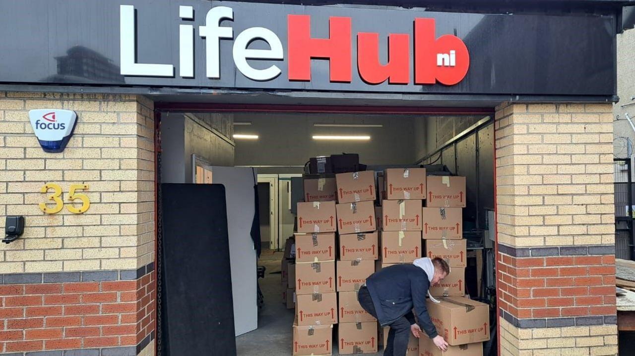 A man lifting cardboard boxes in his hoody and navy jacket. 
The building behind has a black and red sign called 'LifeHub' and it is yellow and red brick. 