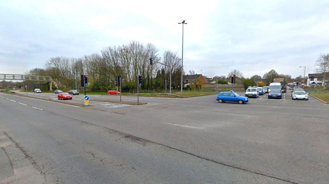 A Google Street View screenshot showing a large junction with three lines of traffic queuing at traffic lights. There are not many cars on the main road and there are trees in the background with very few leaves on them.