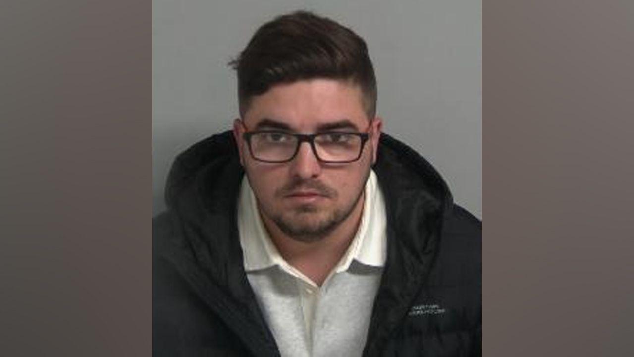 A custody mugshot of Elliot Wright, who is wearing a black coat, grey collared jumper and black rimmed glasses. He has brown hair and a stubble-like beard. He is staring at the camera with a serious expression.