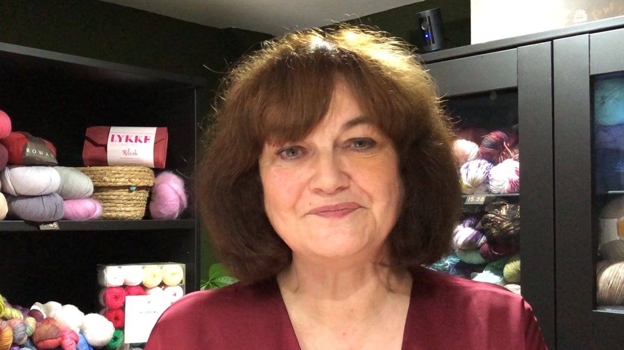 A general view of Sharon Cane, owner of the Knitters Attic. She is standing in the shop and smiling to the camera. She has shoulder length brown hair and is wearing a dark red dress. Behind her are black shelves which have tens of yarn balls placed upon then.