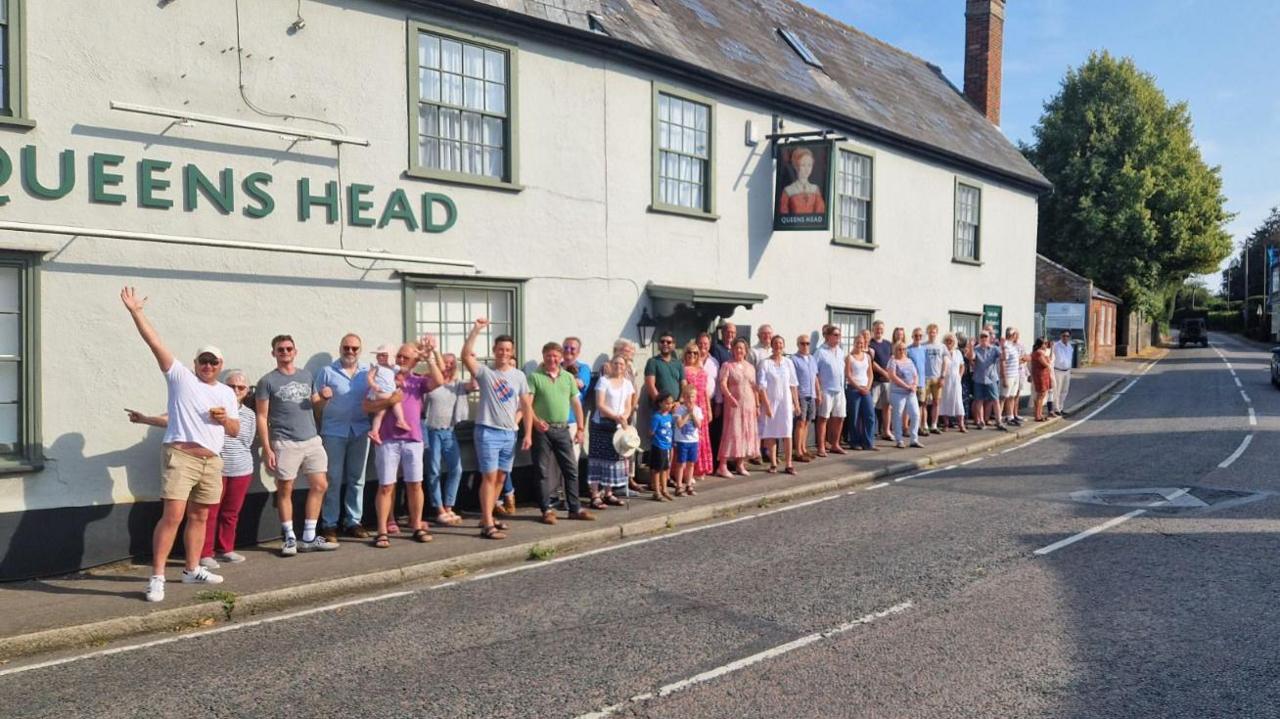 A group of over 30 people stand in front of a white pub with the Queens Head lettering and sign hanging above them