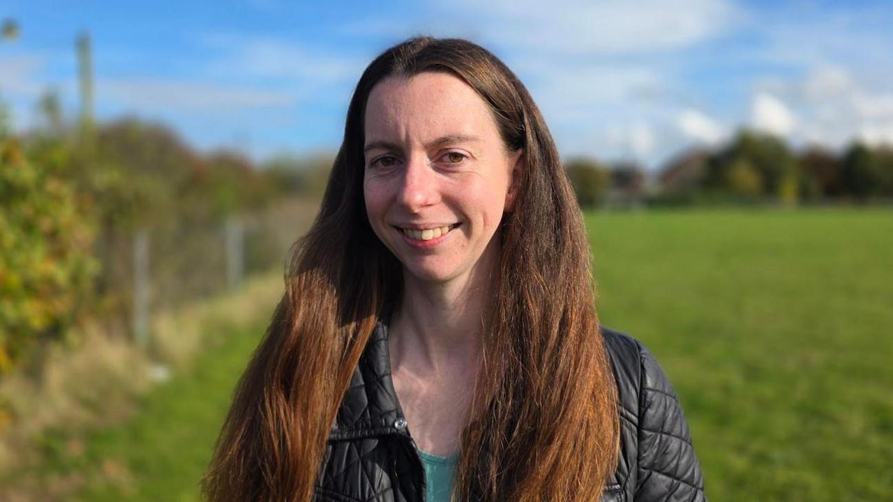 Jess Royal is standing in a field. She's wearing a black jacket over a green top.