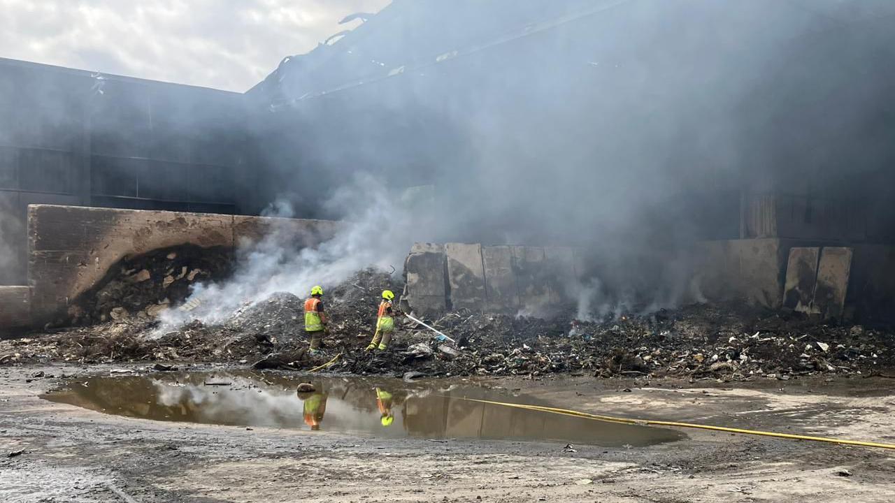 Two fire fighters, standing on chard remains of recycling rubbish, at a large waste site. There is smoke coming up from the rubbish, and concrete structures around the waste.