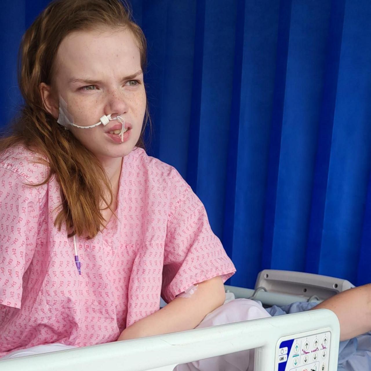 Anastasija is seen with a nasal feeding tube sitting in a hospital bed wearing a pink gown, appearing unwell and looking to the side.