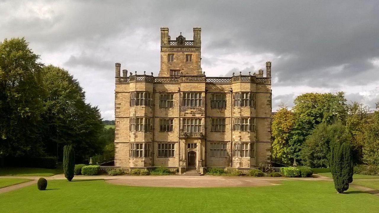 The imposing three-storey stone-fronted Gawthorpe Hall, with a tower in its centre on an additional level. The hall is surrounded by a neatly mown lawn, trees and shrubs.