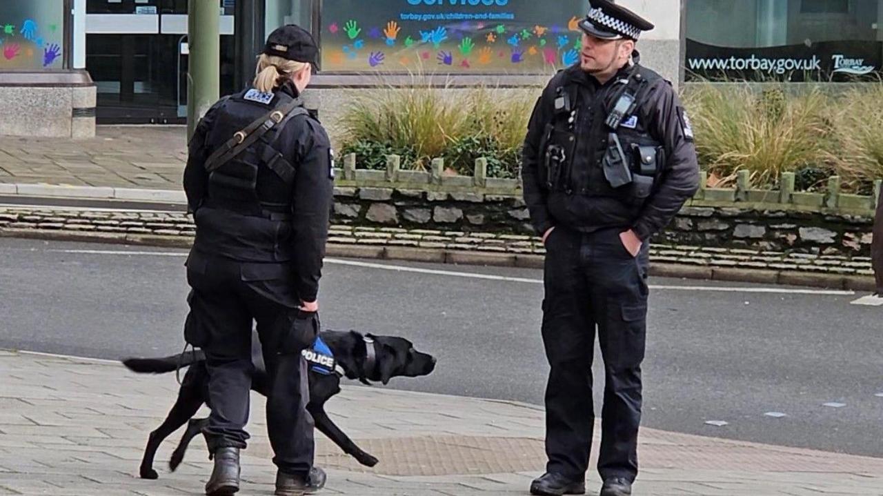 Two police officers in uniform are on a pavement next to a road. The female officer is holding a black Labrador dog on a lead. The dog is wearing a harness that reads POLICE. The male officer is looking a the woman and has is hands in his pockets. Behind them is a shop window decorated with multi-coloured handprint cut-outs and a banner that reads WWW.TORBAY.GOV.UK