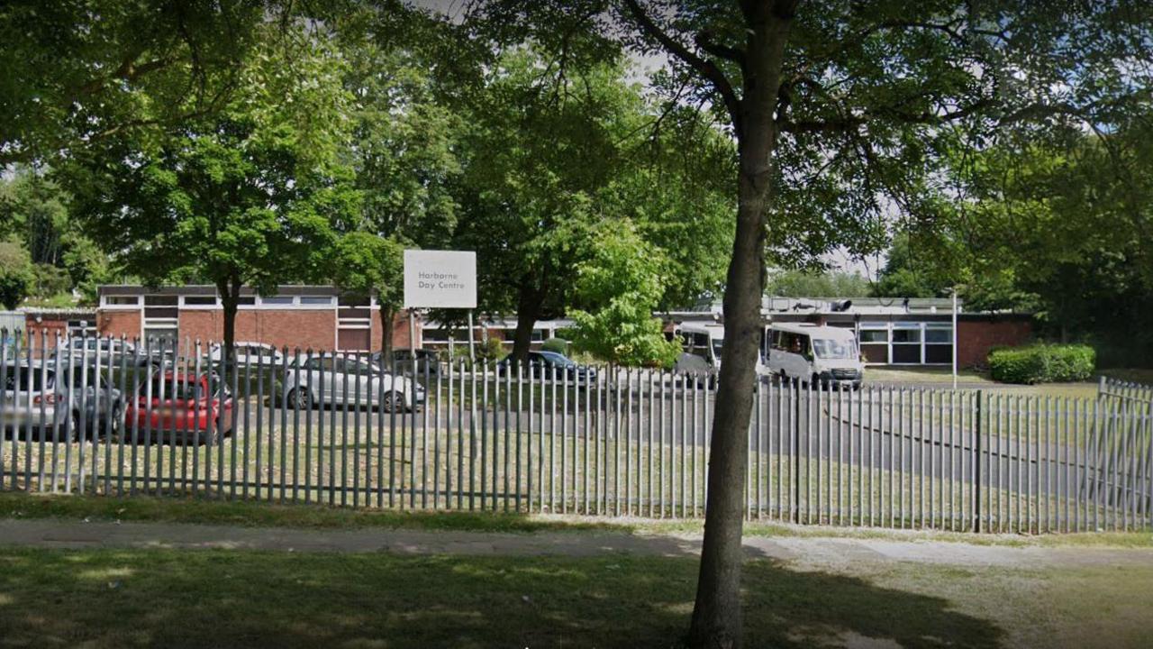 A wide brick building behind metal railings, surrounded by a number of trees, as well as grass and bushes in the background. Around a dozen cars are also parked in the car park in front of the building. 