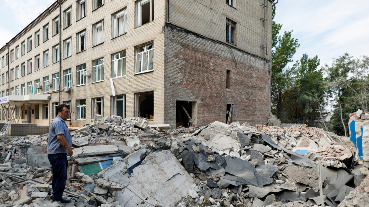 A man in a blue shirt and black trousers standing on the rubble of a fallen building, with another block of flats in the background