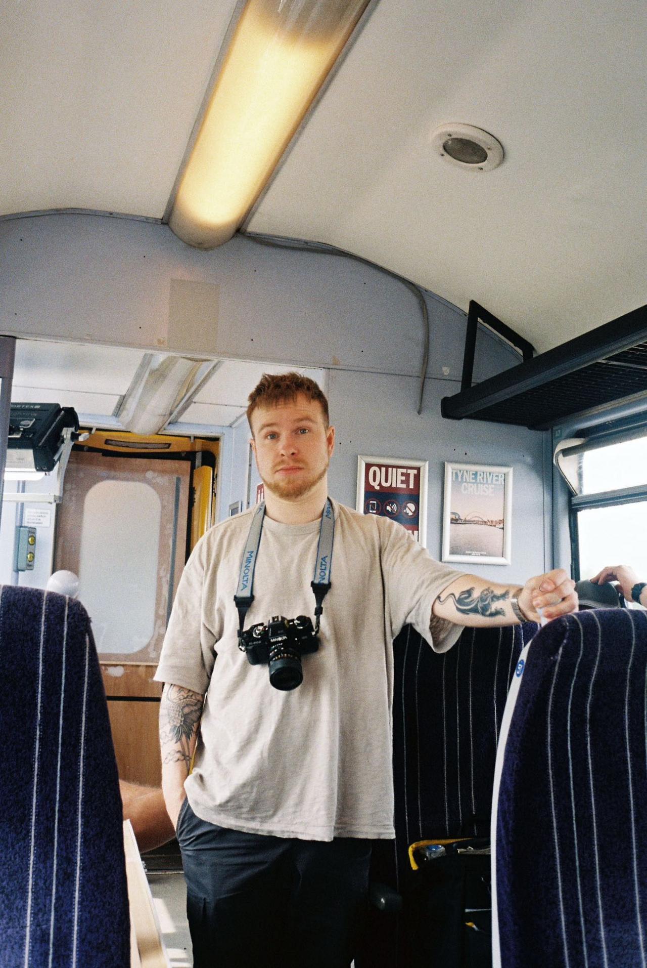 George Dixon stands on a train with a camera slung around his neck. He has short brown hair and a beard, and he is wearing a cream coloured T-shirt. He has tattoos on his arms.