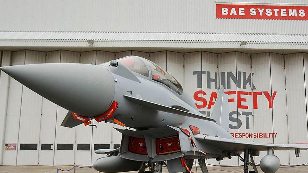 A Eurofighter Typhoon fighter jet at BAE Systems in Warton stands outside a grey warehouse with concertina doors and a red-and-white BAE Systems sign.