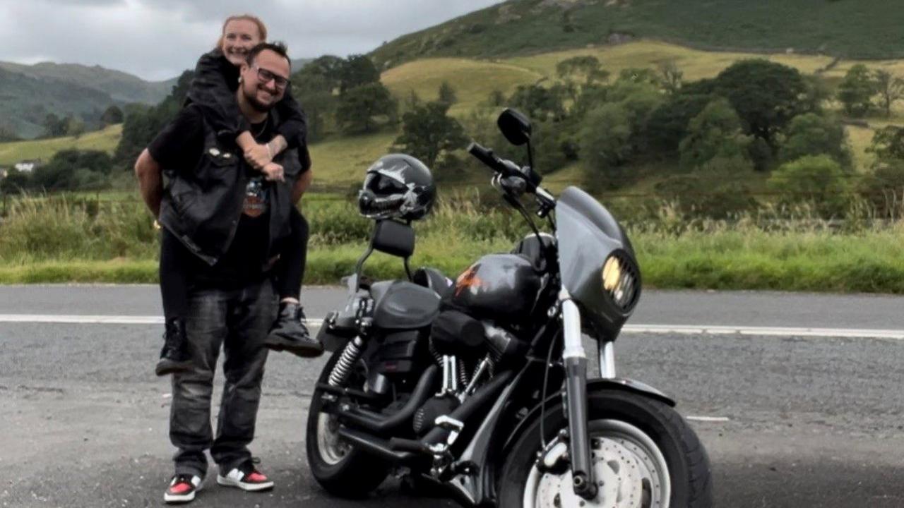 Ryan Crawford gives Laura Burrows a piggyback, with Ryan's motorbike by a road in the Lake District, with a hill in the background