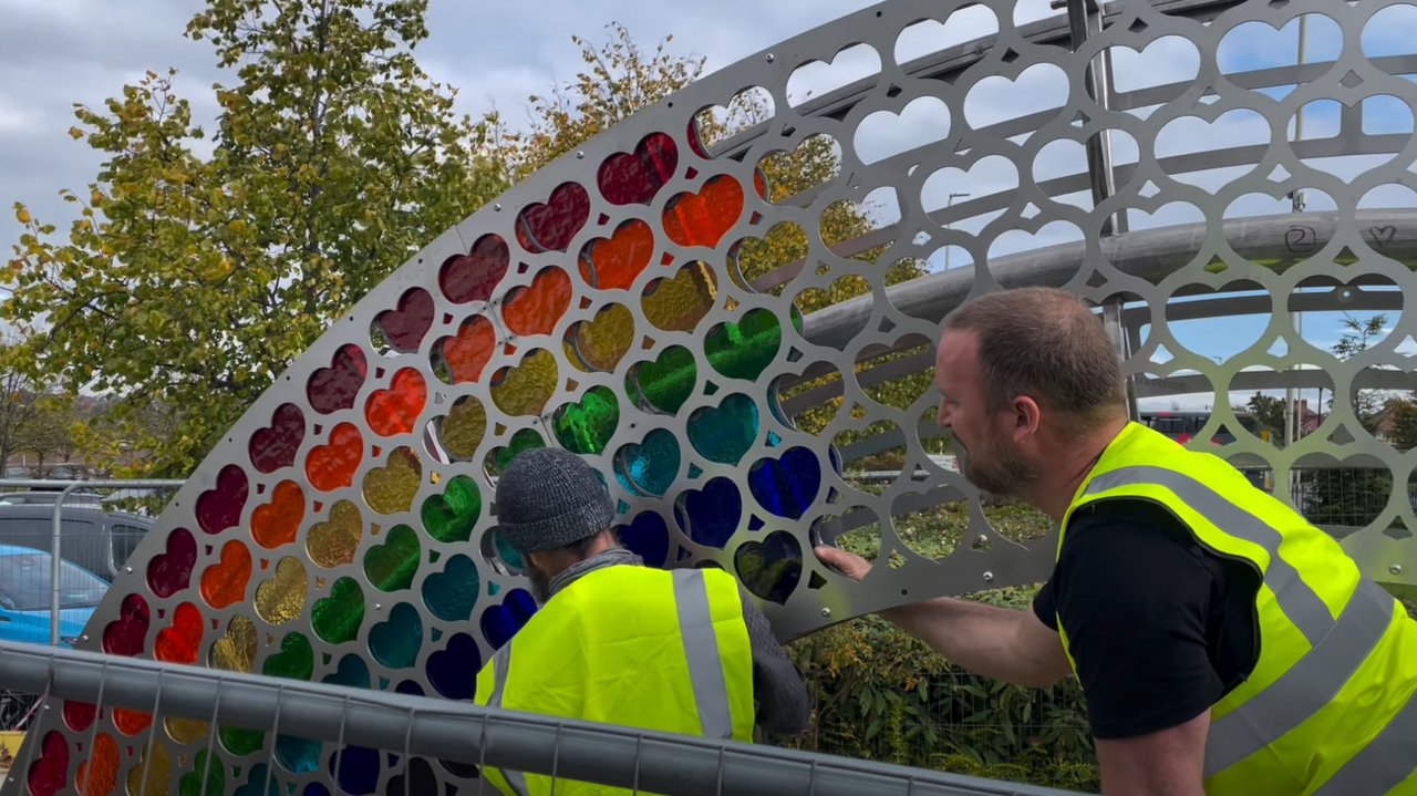 A close-up of two workers wearing hi-viz tabards and work clothes. They are behind security fencing installing glass inserts into heart-shaped holes in a metal rainbow structure.  