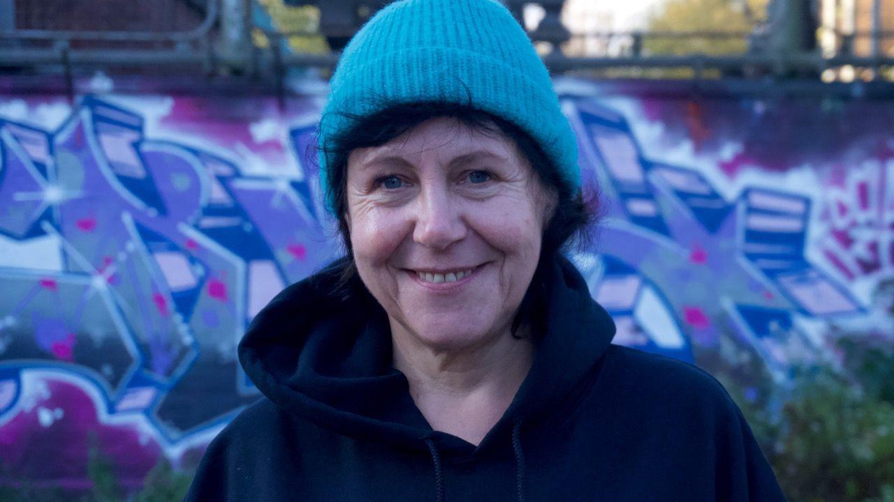 Berni Good is wearing a blue woolly hat and a black jumper. She is standing in front of a wall filled with colourful graffiti