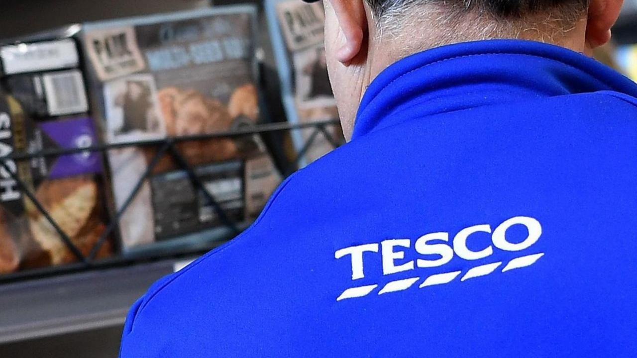 A look over the shoulder of a Tesco employee at a shelf of baked goods which is out of focus. The staff member is wearing blue with "Tesco" written on his back in white font.