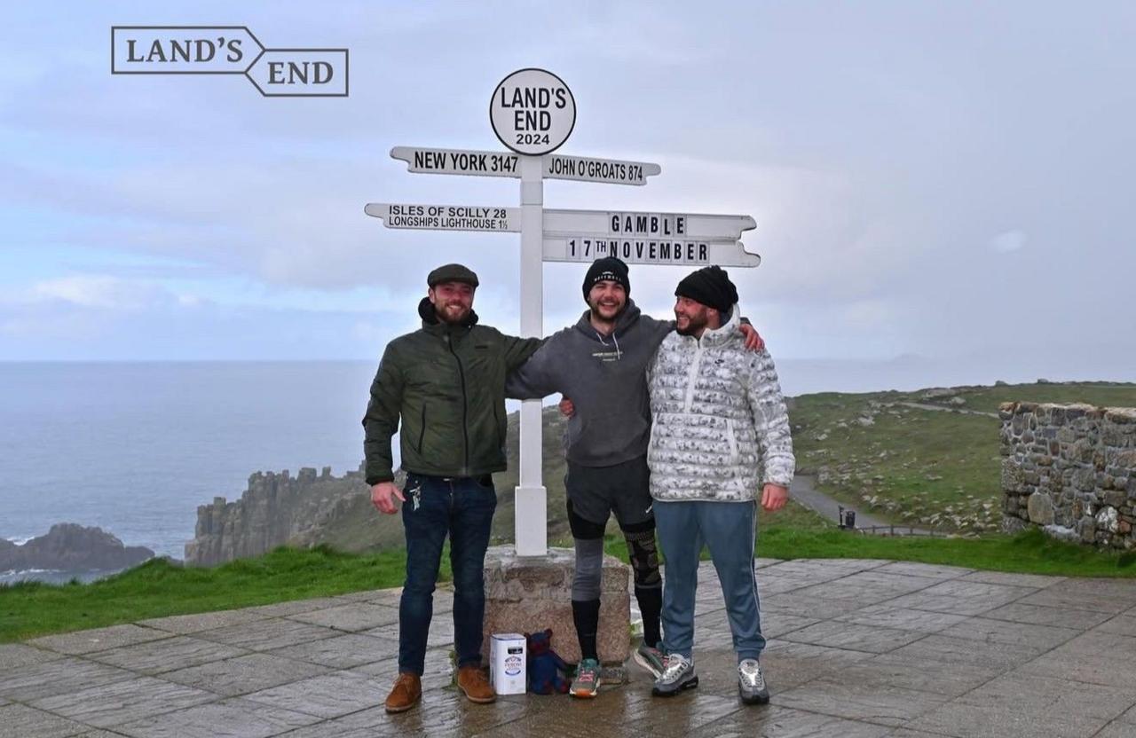 Three people stand in front of a signpost at Land's End in Cornwall, England. The sign displays distances to New York, John O'Groats, Isles of Scilly, and Longships Lighthouse, along with a custom sign reading 'GAMBLE 17TH NOVEMBER'. Cliffs and the ocean are visible in the scenic coastal background.