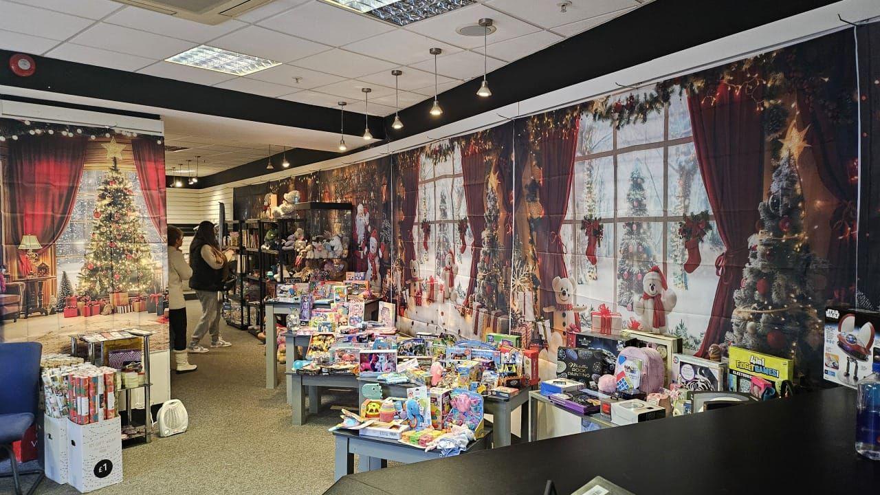 A room filled with toys and games. The walls have been adorned with Christmas decorations and a couple of people are looking at some of the items.