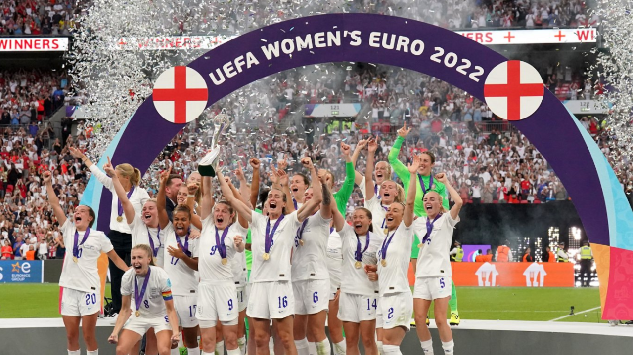 The victorious Lionesses celebrating winning the UEFA Euro 2022 on the pitch, standing in a group wearing their medals, with their hands in the air, and the captain holding the trophy aloft