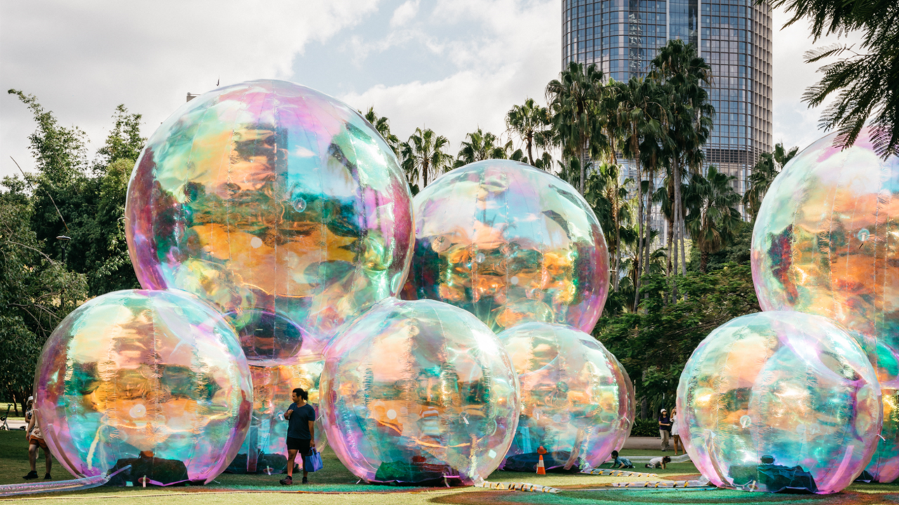 Giant and colourful bubble artwork reflecting from the sunlight. There is a person stood in front of one the instillation. It is at the centre of a park.