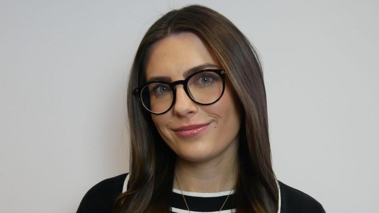 Amy Spooner, who has long brown hair and is wearing glasses with black rims. She is also wearing a black top with white stripes through the middle. She is standing in front of a grey wall.