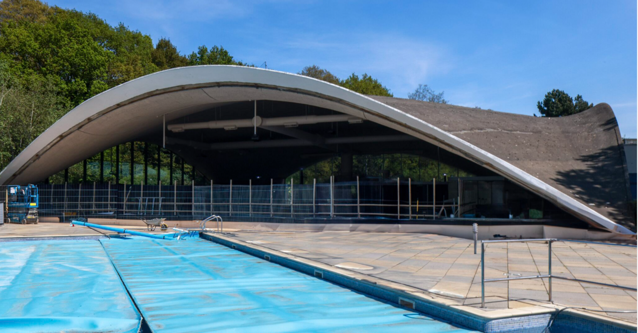 A side view of a concrete shell roof, with a covered outdoor swimming pool in the foreground. 