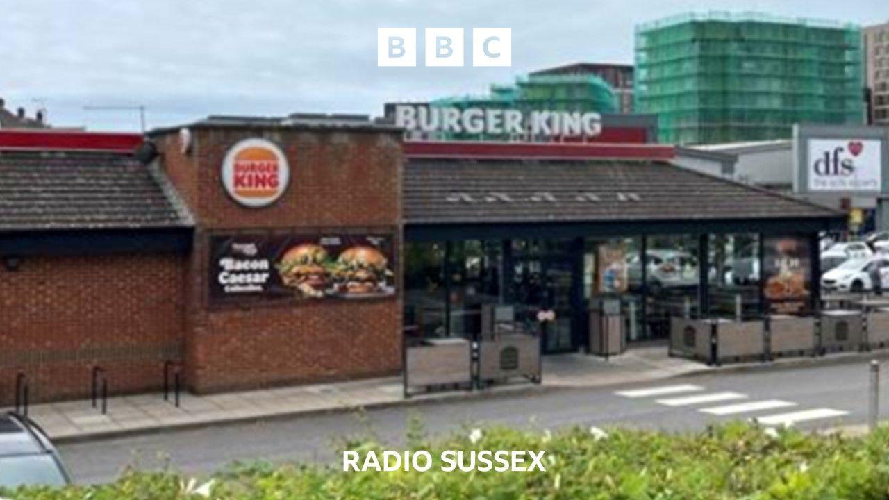 The outside of a single story Burger King restaurant. A poster advertising a Bacon Ceasar burger is displayed on the outside wall. A construction site where flats are surrounded by scaffolding can be seen in the background. 