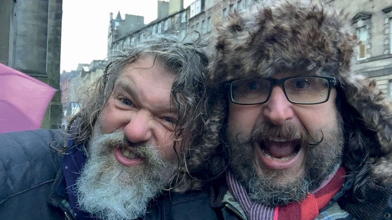 Si and Dave are taking a selfie and making funny faces to the camera. Si has his head resting on Dave's shoulder. Dave is wearing a furry hat and has glasses on. It is cold as they are both wearing winter coats and scarves.