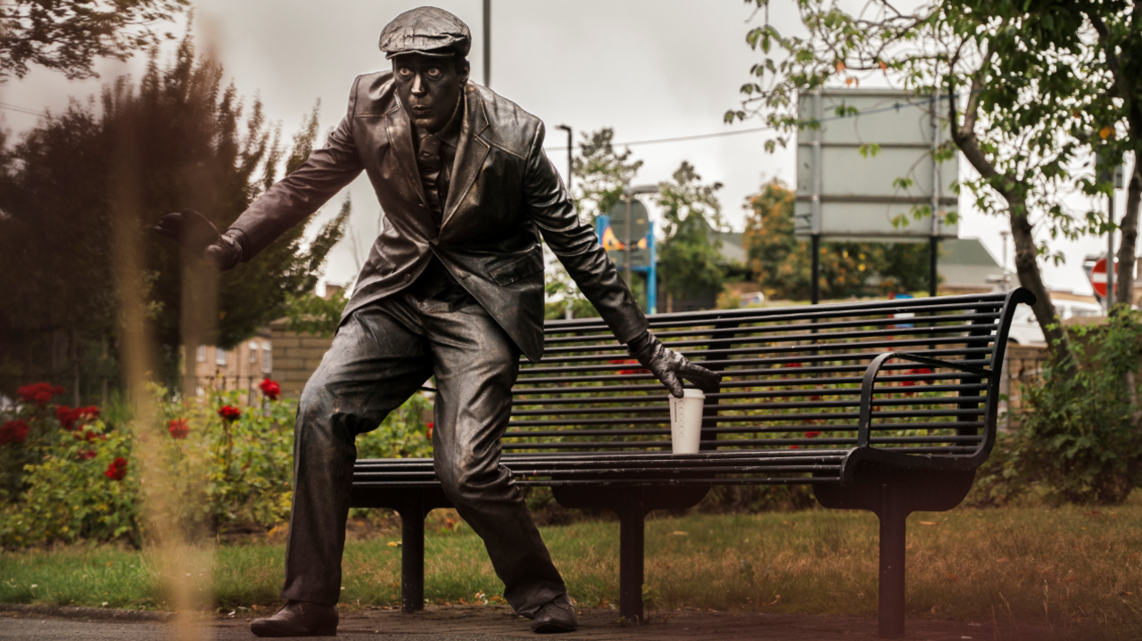 A man wearing a suit and sprayed with metal paint pretends to be a metal statue in a park in Heckmondwike. He is leaving a coffee cup on a bench to highlight "careful littering". Behind him green space is blurred.