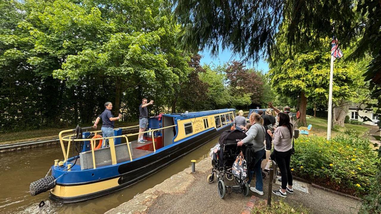 A blue and yellow boat on a canal with a number of people, one of them in a wheelchair, waving from the towpath
