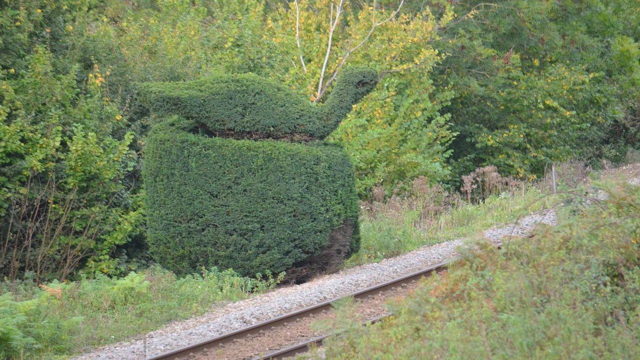 A topiary which is the shape of a bird. It is situated next to a railway line and behind it are trees.