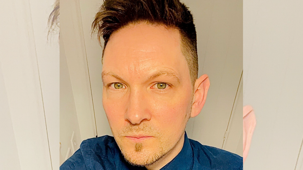 John Payne takes a selfie from a raised angle. He is wearing a blue shirt and is standing in front of a white door. He has green eyes and dark hair, styled in a quiff. 