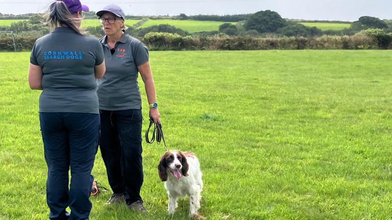 Ms Page has her back to the camera as she talks to Ms Simpson who has springer spaniel Freda on a lead standing at her feet. They are in a grassy field and both are wearing the Cornwall Search Dogs branded T-shirts with trousers.