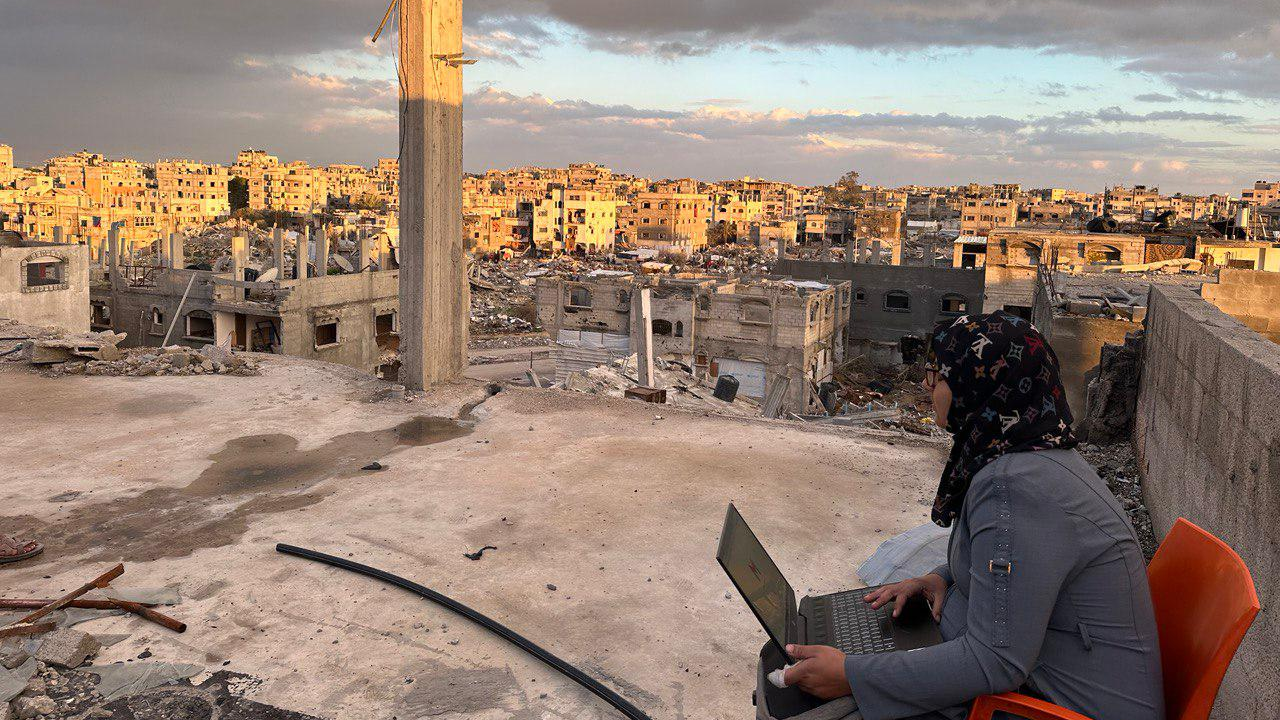 Manar sits on an orange chair with a laptop on her lap. She is looking out into the distance and wearing a dark blue jacket. In the background are buildings falling apart and with roofs off and various bits of rubble.