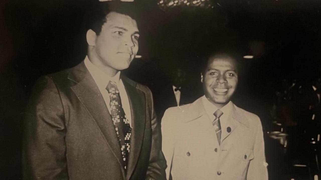 Dr Stephenson (right) pictured with Muhammad Ali. Dr Stephenson is smiling at the camera in a shirt, tie and light coat, and appears to be walking with Mr Ali who is wearing a tie and dark jacket, and is smiling at a different camera. 