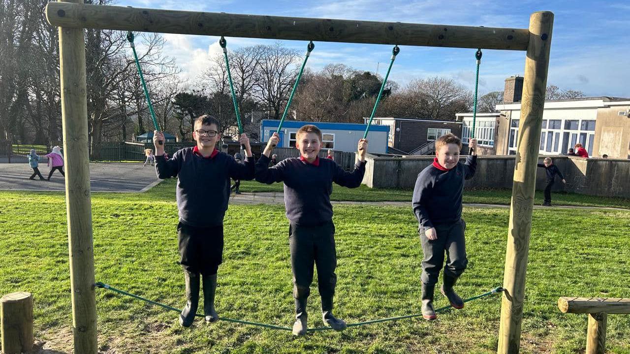 Three primary school boys stand on a rope balancing beam in a playing field while holding on to rope handles, with the school behind them.